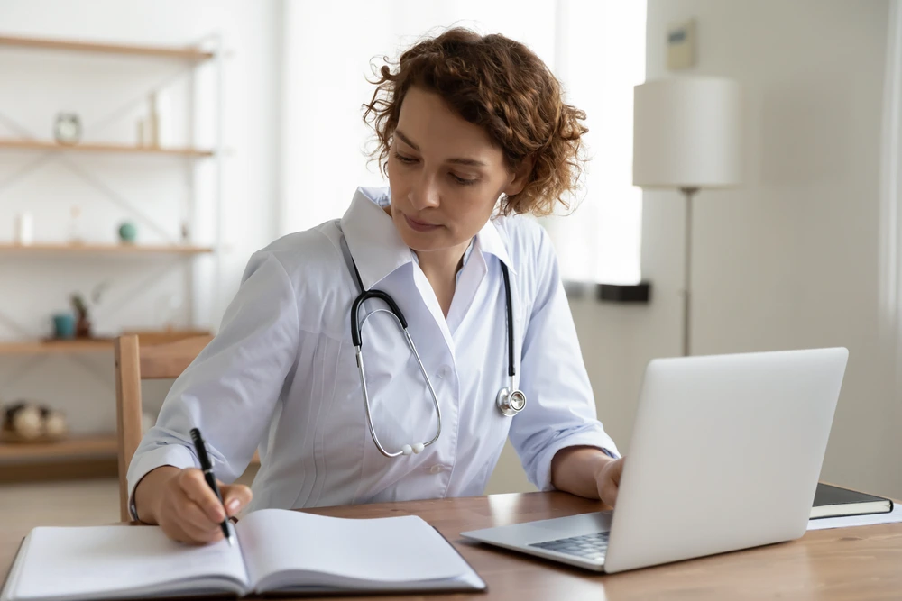 serious-female-doctor-using-laptop-writing-notes-in-medical-journal