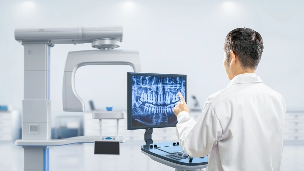 A dentist reviewing a panoramic dental X-ray on a monitor next to a dental imaging machine.