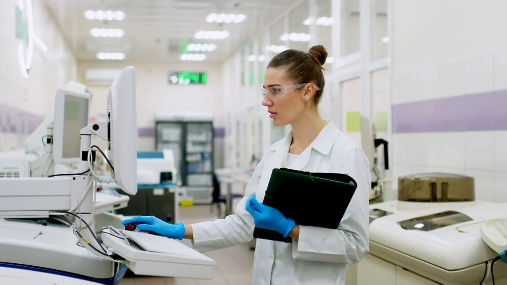 A female scientist in a lab coat and safety glasses wearing blue gloves, working at a computer in a laboratory setting, holding a black folder.