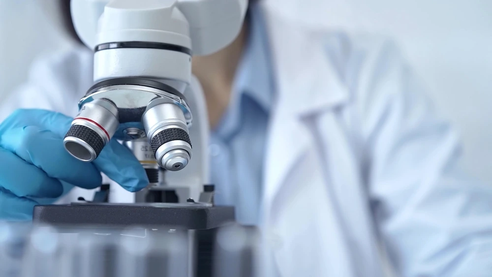 A close-up of a person in a lab coat and blue gloves adjusting a microscope.